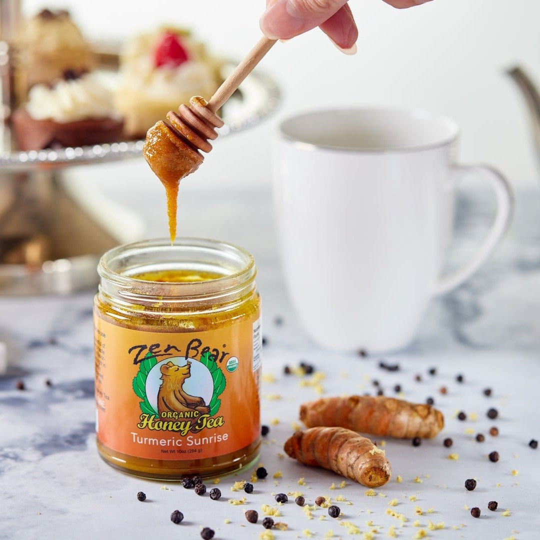 Jar of honey with a dipper, surrounded by tea and pastries on a table.