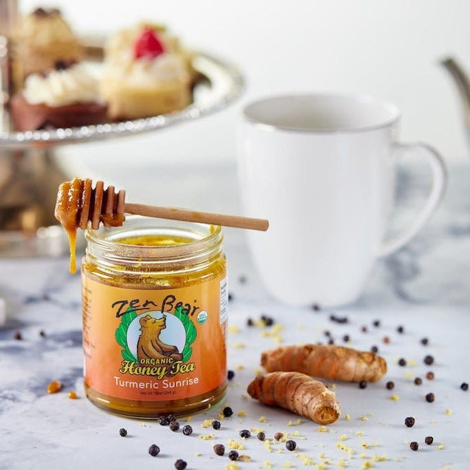Jar of honey with a dipper on a table with tea and pastries in the background
