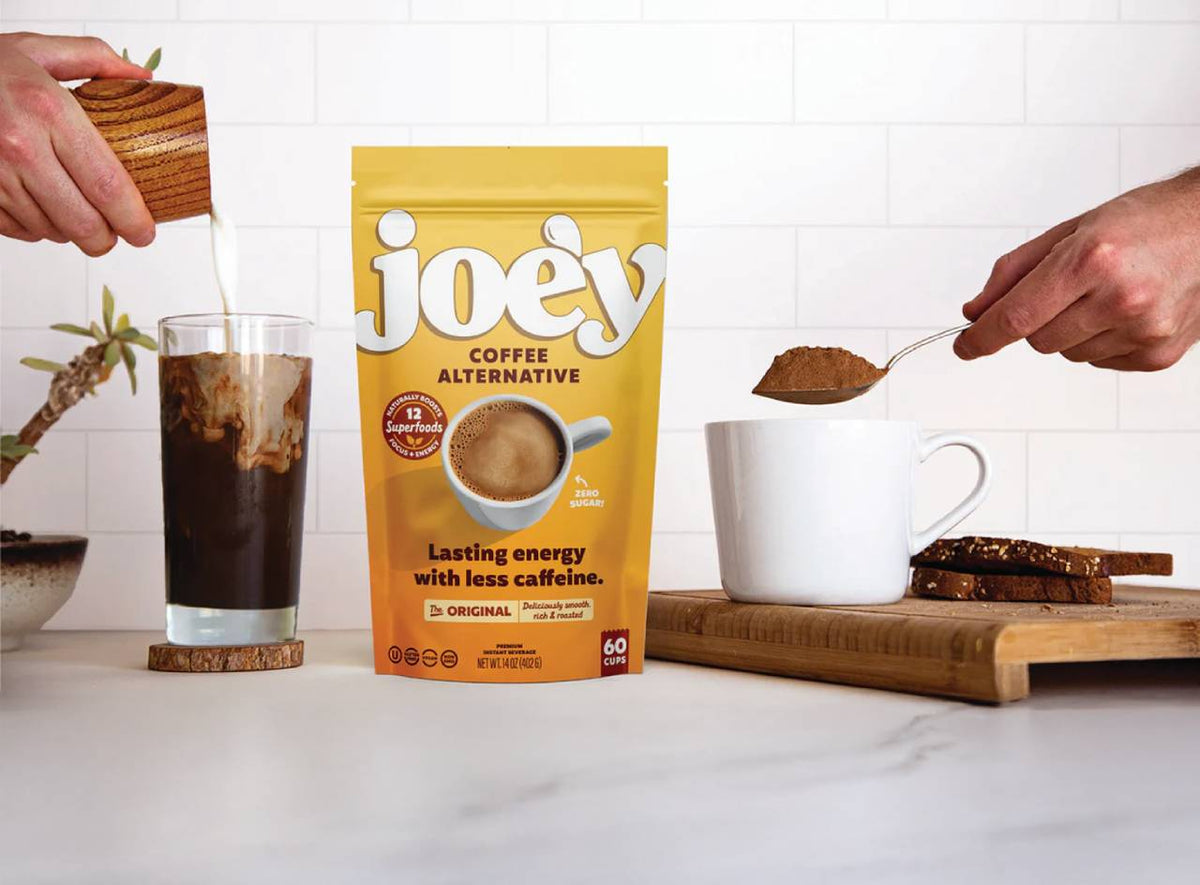 A stand-up pouch of Joey Coffee Alternative is centered on a countertop, with hands shown preparing coffee around it: pouring milk into an iced coffee glass on the left and adding powder to a white mug on the right. The package is yellow-orange with large white &