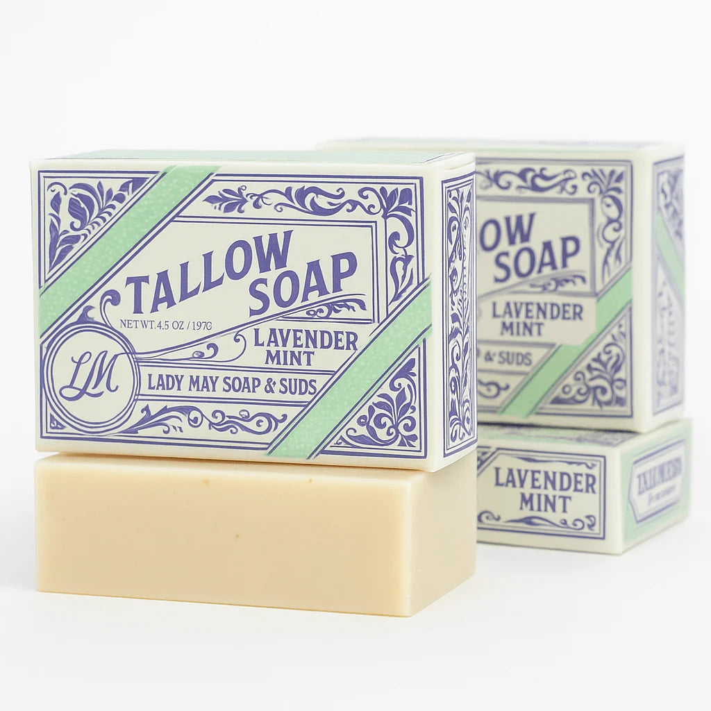A bar of Tallow Soap, Lavender Mint, stacked on top of two matching boxes in the background. The soap bar is a warm cream color, and the boxes feature blue and light green decorative labels with the brand name &