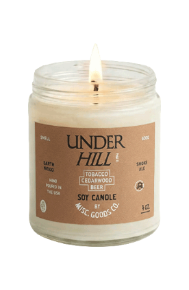 A lit 7 oz soy candle in a clear glass jar with a brown paper label. The label reads &