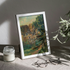 A framed landscape art print with a white frame, leaning on a draped fabric surface next to a candle jar, eyeglasses, and a vase with green leaves. The scene is lit by dramatic, diagonal natural sunlight casting shadows on a textured gray wall.