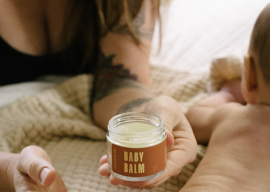 Person holding a jar of baby balm next to a baby on a soft surface
