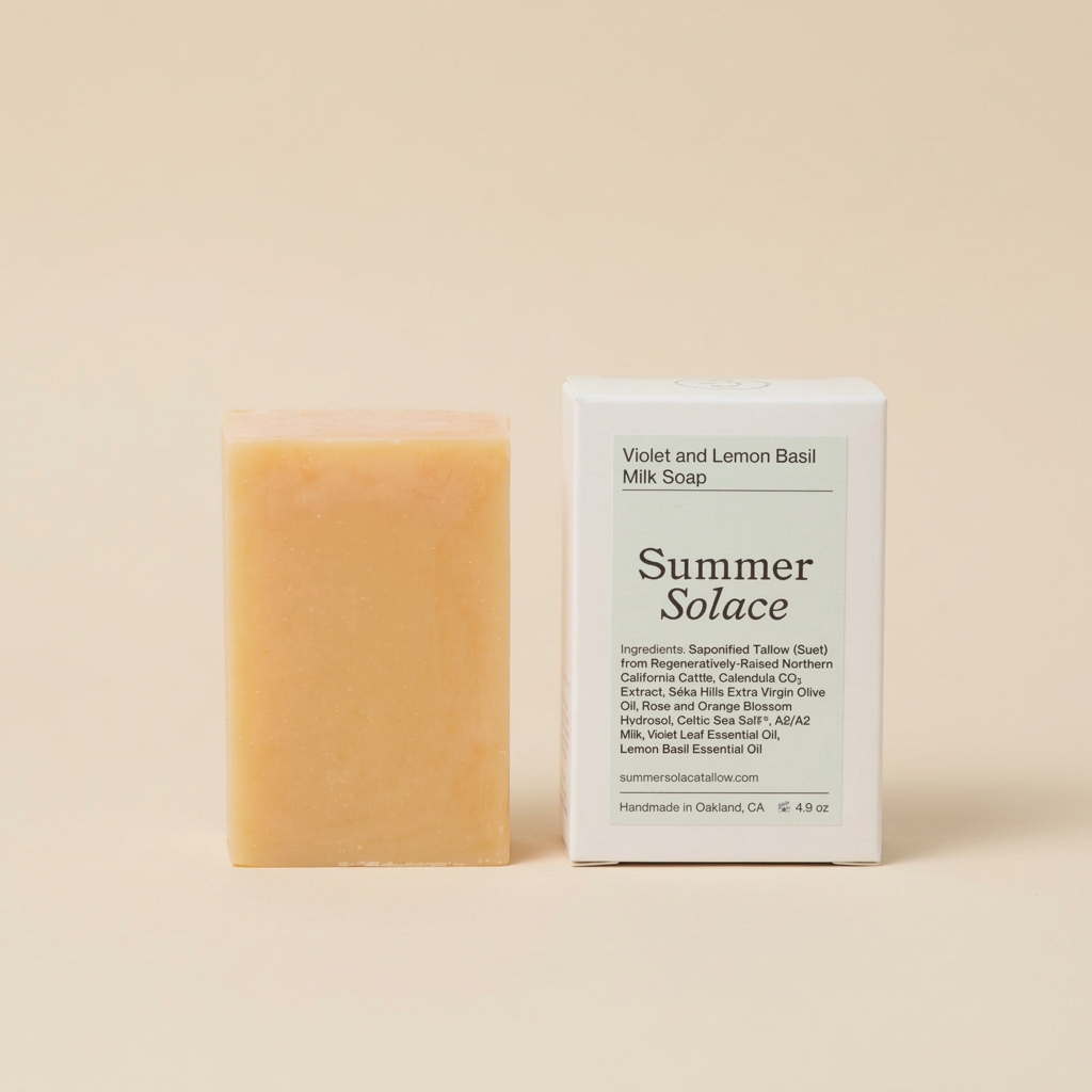 A rectangular bar of pale yellow soap stands beside its white product box on a warm beige studio background. The box reads &