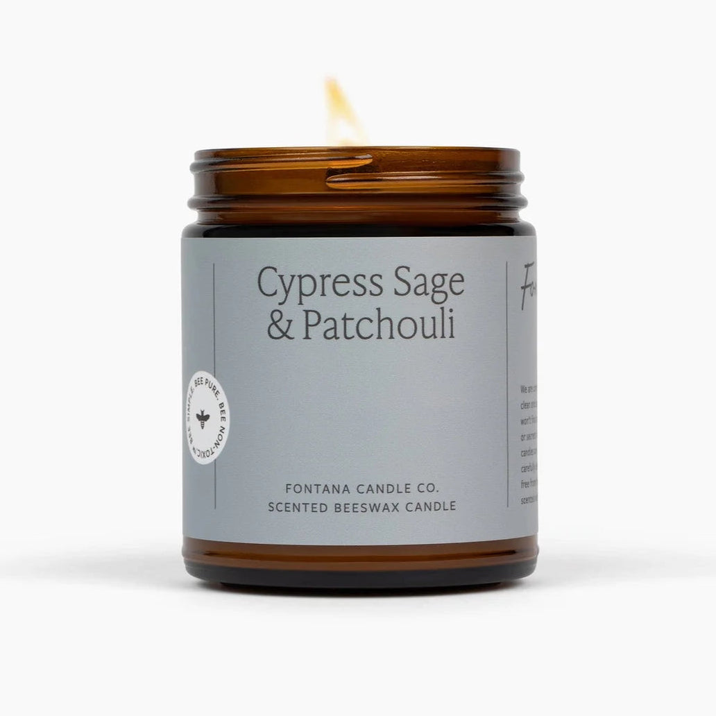 A lit amber glass jar candle with a gray label reading &