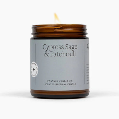 A lit amber glass jar candle with a gray label reading &
