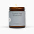 A lit amber glass jar candle with a gray label reading &