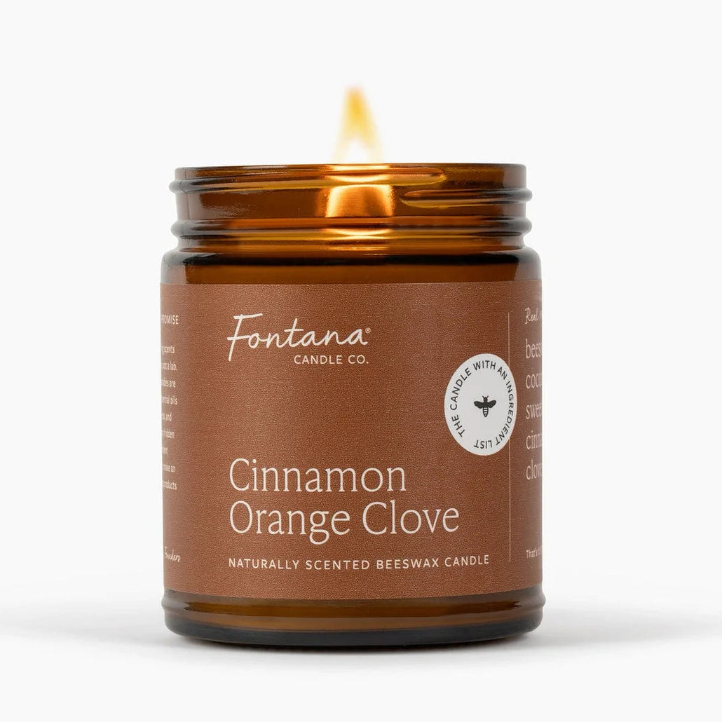 A lit Fontana Candle Co. Cinnamon Orange Clove scented beeswax candle in an amber glass jar with a brown label, set against a clean white background.
