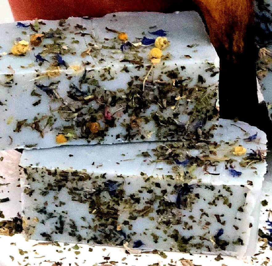 Two stacked rectangular bars of handmade soap with a white base, heavily speckled with dried green herbs, and decorated with small dried flowers in yellow, blue, and orange, set against a rustic background.