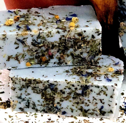 Two stacked rectangular bars of handmade soap with a white base, heavily speckled with dried green herbs, and decorated with small dried flowers in yellow, blue, and orange, set against a rustic background.
